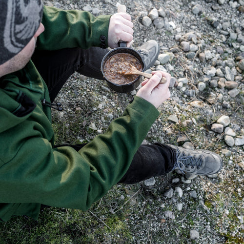 Person dressed in green jacket seated on rocky ground stirring food in a black enamel pan with a wooden handle, perfect for camping, kitchen, cookware, and outdoor use collections.