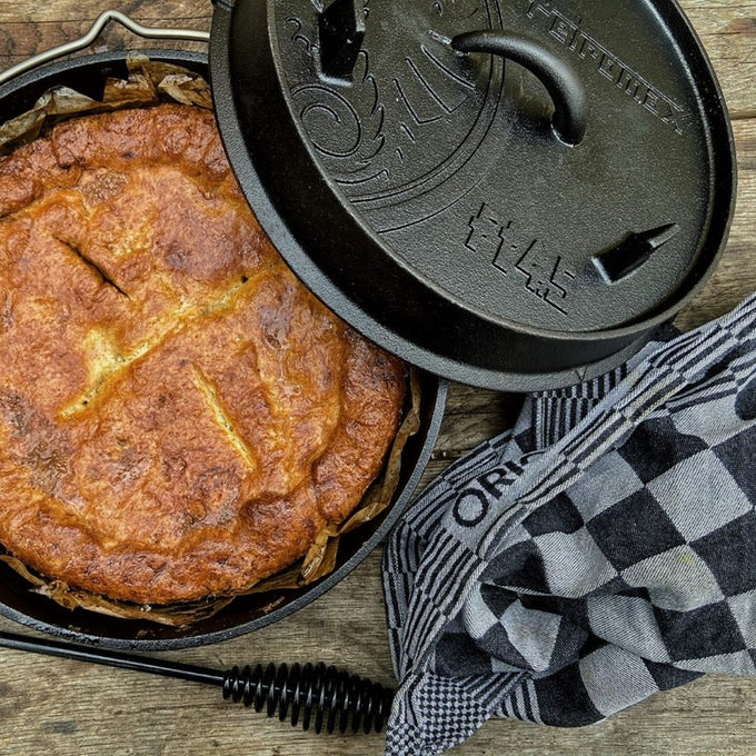Black cast iron Dutch oven with legs displaying a freshly baked golden pie inside, accompanied by a checkered kitchen towel, perfect for Kitchen & Food, Cookware, Cast Iron collections.