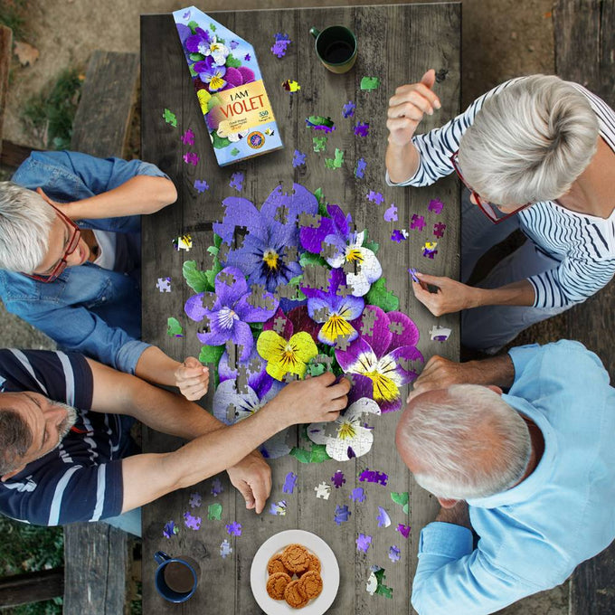Four adults assembling a unique-shaped violet-themed jigsaw puzzle on a wooden table outdoors, surrounded by puzzle pieces, coffee mugs, and a plate of cookies, part of Toys, Books & Games collection.