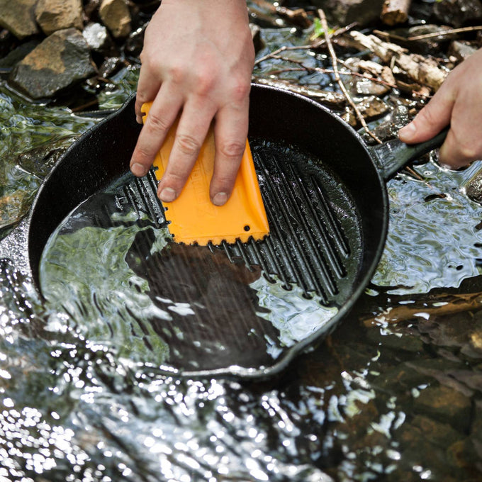 Hands using an orange pan scraper to clean a cast iron grill pan submerged in flowing water, highlighting durable cookware care in Kitchen & Food and Cast Iron collections.