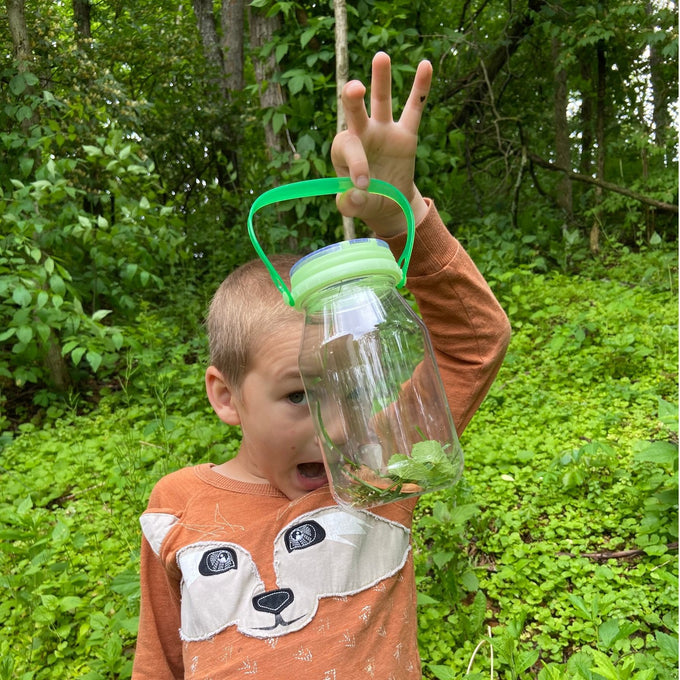 Child outdoors holding a handcrafted clear plastic bug catcher with a green glow-in-the-dark magnifying lid and handle, surrounded by green foliage in a natural setting, promoting outdoor play and exploration from the Toys, Books & Games and USA Made collections.