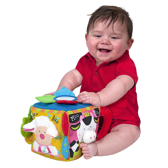 Baby in red outfit playing with colorful fabric musical farmyard cube featuring animal faces, textured details, and interactive sound elements, ideal for indoor play and learning.