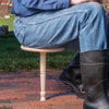 Man wearing denim jeans and black rubber boots sitting on a handcrafted hardwood one-legged stool outdoors on brick flooring, featured in Farm, Garden & Tool, Animal Care, USA Made, and Best Sellers collections.