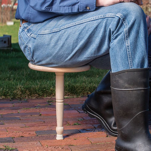 Man wearing denim jeans and black rubber boots sitting on a handcrafted hardwood one-legged stool outdoors on brick flooring, featured in Farm, Garden & Tool, Animal Care, USA Made, and Best Sellers collections.