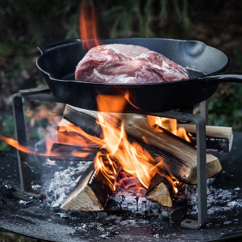 Cast iron skillet with long handle cooking a thick raw meat cut over an open wood fire on a sturdy metal tripod, ideal for Farm, Garden & Tool, Kitchen & Food, Camping, Cookware, Cast Iron, Best Sellers, and Promo Eligible collections.