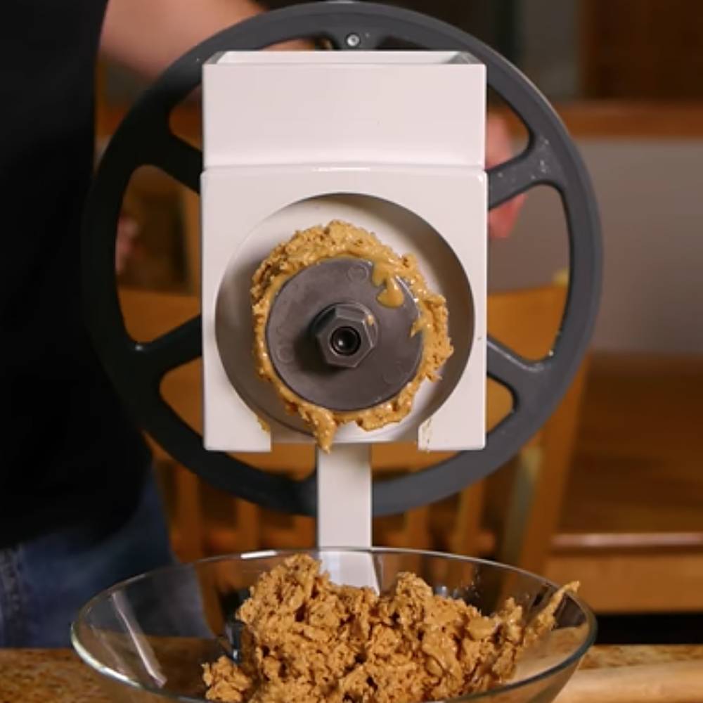 Close-up of a nut butter accessory attached to a grain mill, showing freshly ground peanut butter being produced into a glass bowl, highlighting a handcrafted, USA-made kitchen tool ideal for food and bread making supplies.