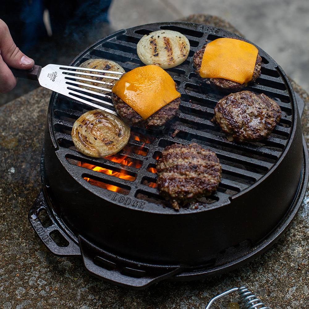 Cast iron portable round grill with burger patties topped with melted cheddar cheese and sliced onions cooking over an open flame, ideal for camping and outdoor grilling.