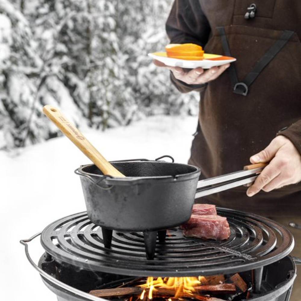 Black cast iron Dutch oven with legs cooking over an open fire grill outdoors, accompanied by wooden utensils and raw steak, ideal for Kitchen & Food, Cookware, Cast Iron, Best Sellers, and Promo Eligible collections.