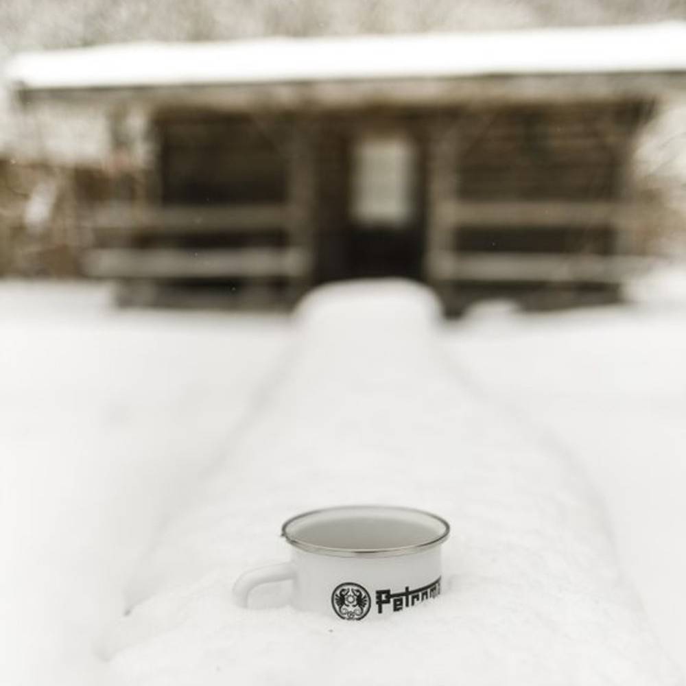 White enameled steel mug with metal rim and black logo, resting on a snow-covered surface in front of a rustic wooden cabin, ideal for Farm, Garden & Tool, Camping, and Brewing Coffee and Tea collections.