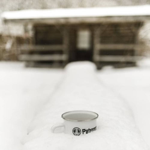 White enameled steel mug with metal rim and black logo, resting on a snow-covered surface in front of a rustic wooden cabin, ideal for Farm, Garden & Tool, Camping, and Brewing Coffee and Tea collections.