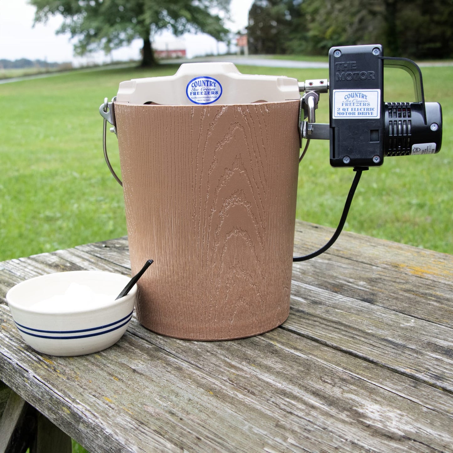 Amish-made electric ice cream freezer with a poly-insulated bucket and attached motor, shown outdoors on a weathered wooden table beside a ceramic bowl, featured in Kitchen & Food and Specialty Equipment collections.