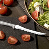 Serrated blade tomato slicer with a polished aluminum handle, displayed next to freshly cut tomato wedges and a bowl of salad with leafy greens and feta, handcrafted in USA for kitchen and food prep collections.