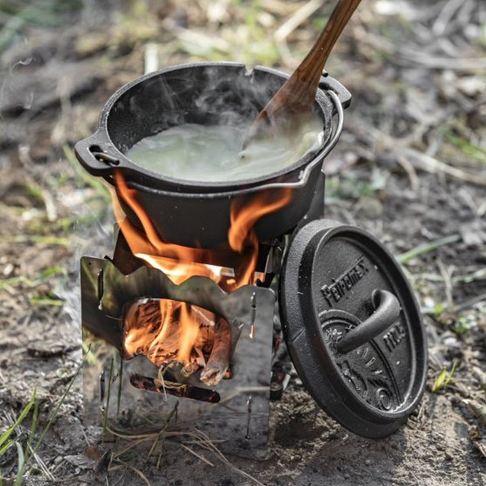 Outdoor cooking scene showing a compact stainless steel hobo stove with a burning wood fire, supporting a cast iron pot with steam rising, suitable for Camping, Farm, Garden & Tool, and Emergency Supplies collections.