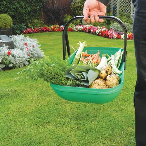 Handheld green plastic colander trug with black carry handles filled with freshly harvested vegetables, standing on a lawn with garden flowers in the background, featured in Farm, Garden & Tool and Garden Accessories collections.