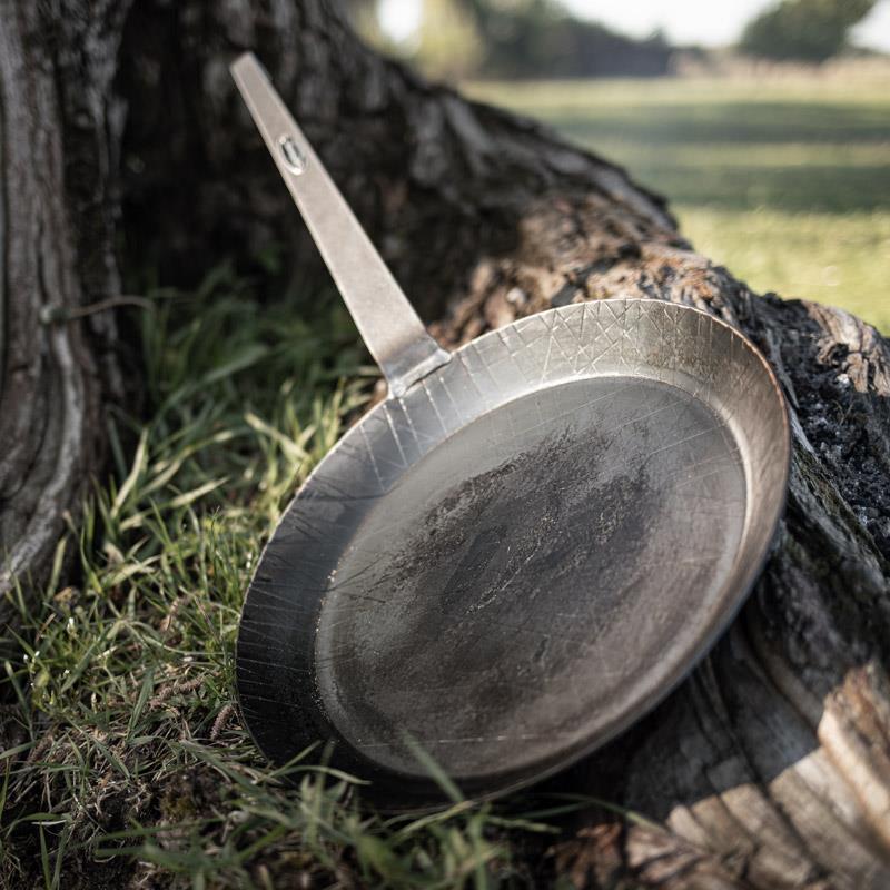 Handcrafted wrought iron pan with a long handle resting against a tree trunk outdoors, showcasing durable construction for Farm, Garden & Tool, Kitchen & Food, Camping, Cookware, and Best Sellers collections.
