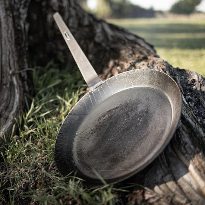 Handcrafted wrought iron pan with a long handle resting against a tree trunk outdoors, showcasing durable construction for Farm, Garden & Tool, Kitchen & Food, Camping, Cookware, and Best Sellers collections.