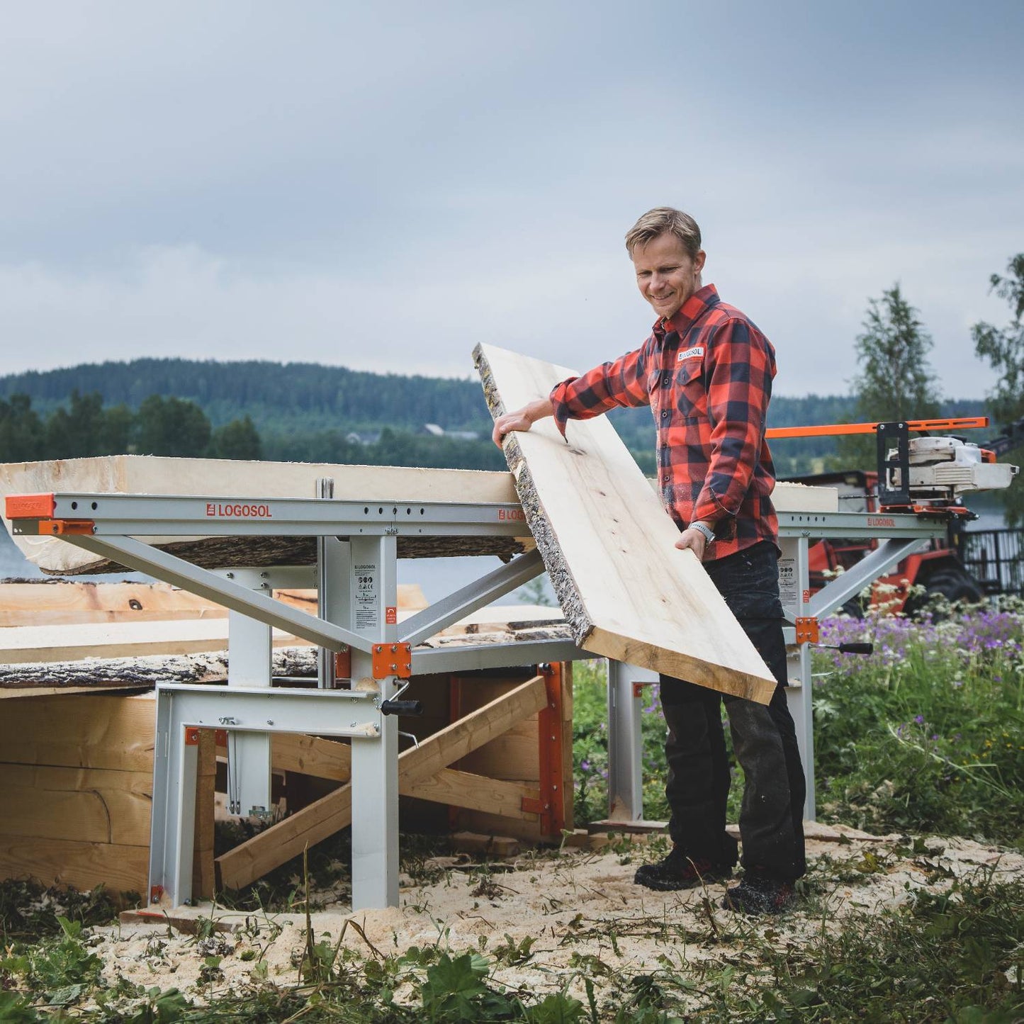 Man in a red plaid shirt lifting freshly cut lumber from a portable silver and orange chain sawmill outdoors with forested hills in the background, suited for Farm, Garden & Tool, Wood Cutting and Hauling, and Best Sellers collections.