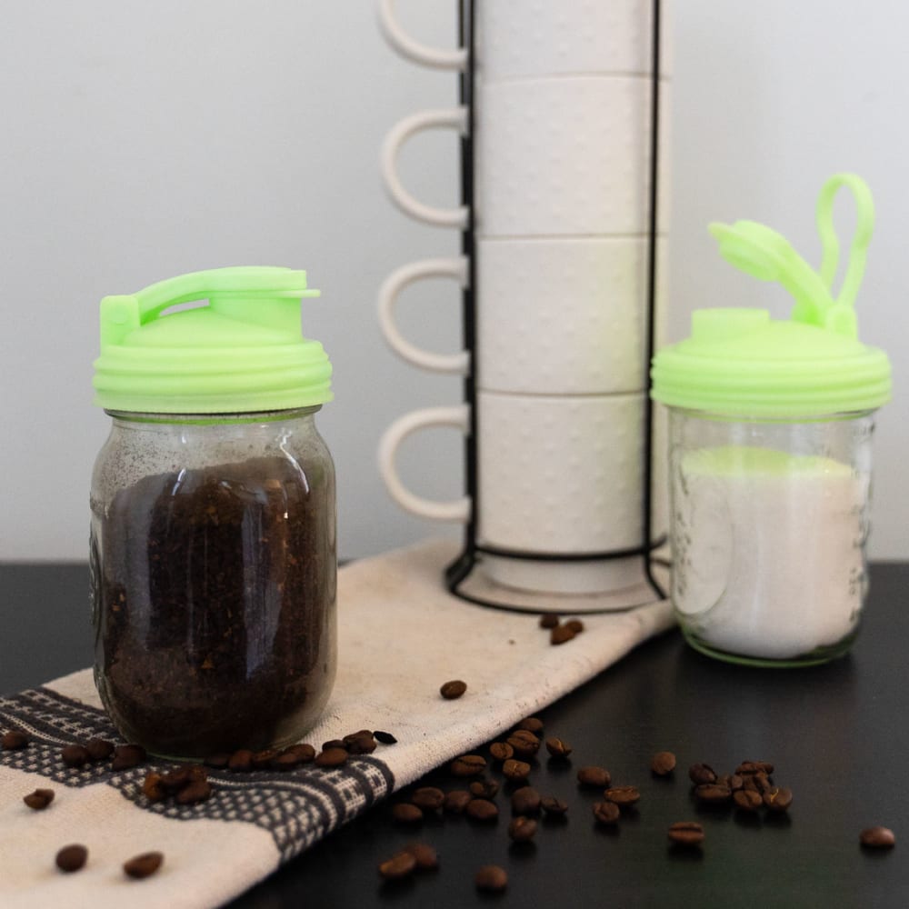 Two glass wide-mouth jars with bright green pour lids featuring flip-top spouts and carry loops, one filled with coffee grounds, the other with sugar, on a kitchen counter with scattered coffee beans, promoting USA Made Kitchen & Food and Best Sellers collections.
