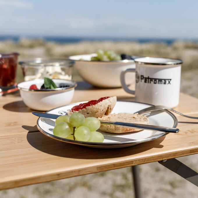Two lightweight enamel plates with black rims and logo, one holding a bread roll with jam and green grapes, paired with matching enamel mug and bowls on a wooden table outdoors, perfect for Farm, Garden & Tool, Kitchen & Food, Camping, Dinnerware, Best Sellers, and Promo Eligible collections.