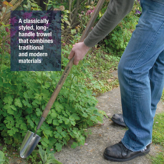Gardener using a stainless steel long-handled trowel with a weatherproofed wooden handle to turn soil between plants in a garden bed, part of farm and lawncare tools.