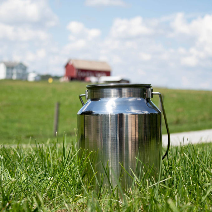 Shining 304 grade stainless steel milk can with seamless construction and sturdy handle, sitting on green grass with a red barn and farmhouse in the background, ideal for farm and animal care use.