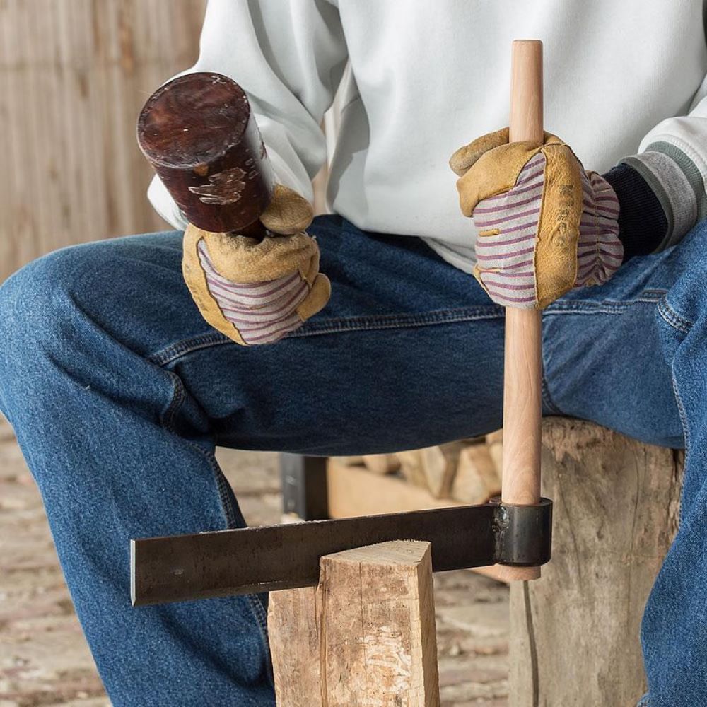 Close-up of gloved hands holding a wooden mallet and a handcrafted wood splitting froe with a 16" hickory handle and case-hardened steel blade, used for wood cutting and hauling in farm and garden tasks.