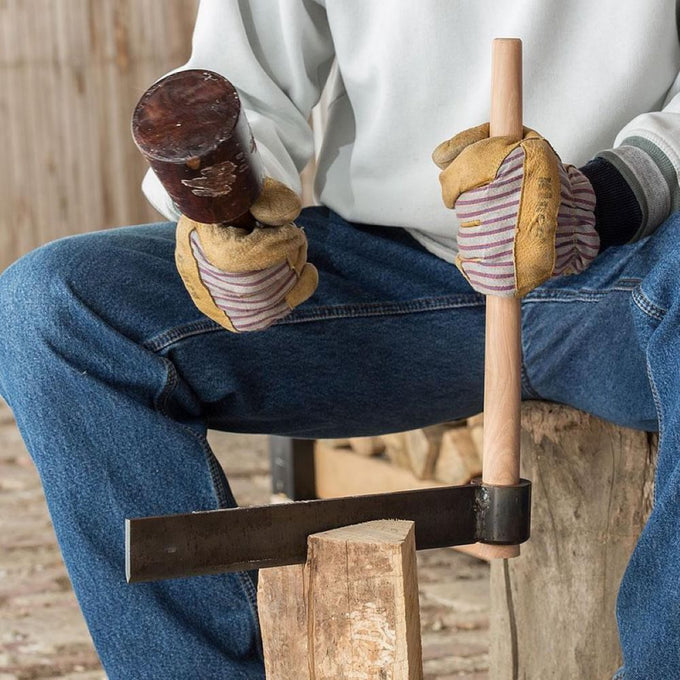 Close-up of gloved hands holding a wooden mallet and a handcrafted wood splitting froe with a 16" hickory handle and case-hardened steel blade, used for wood cutting and hauling in farm and garden tasks.