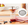Stainless steel funnel with removable mesh filter set on a glass jar containing red liquid, surrounded by blueberries, wooden spoon, and kitchen towel in a bright kitchen setting.