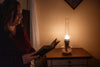 Woman reading a book by the warm glow of a traditional non-electric oil lamp on a wooden side table, evoking a cozy home and garden how-to book setting.