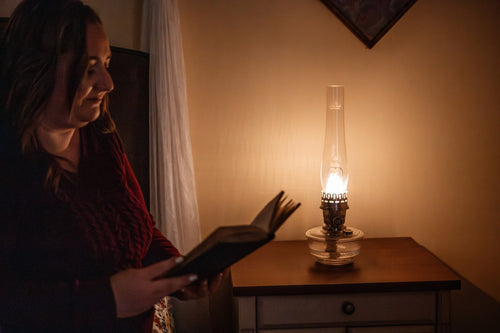 Woman reading a book by the warm glow of a traditional non-electric oil lamp on a wooden side table, evoking a cozy home and garden how-to book setting.