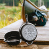 Two seasoned cast iron skillets with one lid featuring a cozy cabin scene, resting on a weathered wooden dock beside camping gear and a canoe, highlighting rugged cookware ideal for outdoor cooking and camping collections.