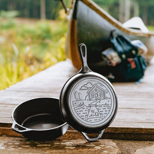 Two seasoned cast iron skillets with one lid featuring a cozy cabin scene, resting on a weathered wooden dock beside camping gear and a canoe, highlighting rugged cookware ideal for outdoor cooking and camping collections.
