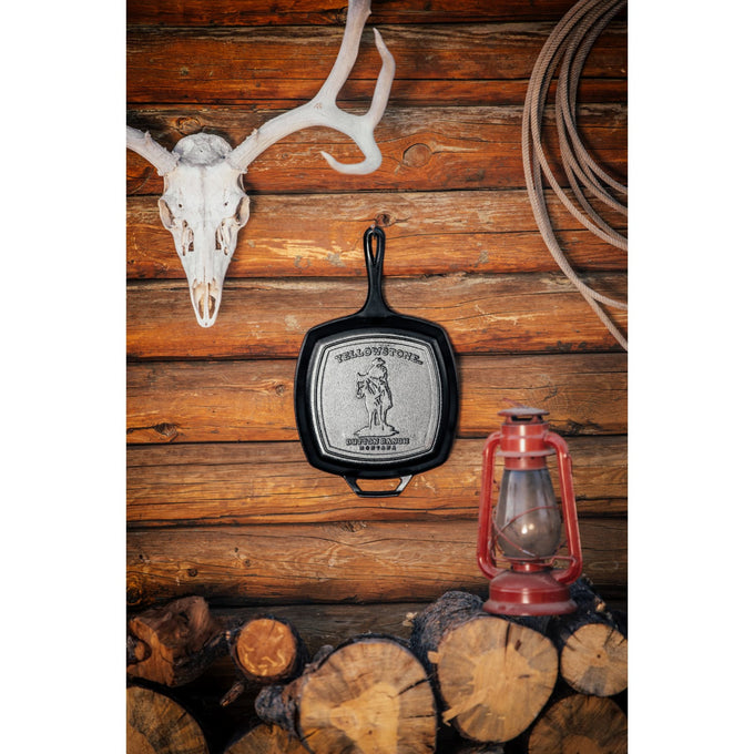Handcrafted cast iron square grill pan with an embossed cowboy design, made in USA, displayed hanging on a rustic log cabin wall with firewood and a vintage lantern, ideal for Kitchen & Food, Cookware, Cast Iron, and USA Made collections.