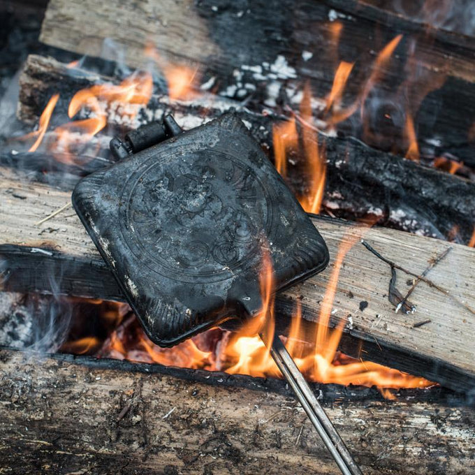 Cast iron sandwich iron with a hinged design and decorative embossed lid, held over open campfire flames and wood, ideal for toasting snacks in Farm, Garden & Tool, Camping, Best Sellers, and Promo Eligible collections.