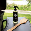 Clear spray bottle of natural BBQ grill oil cleaner with black label and wooden spatula resting on a black grill surface outdoors, showcased for home and grilling use.