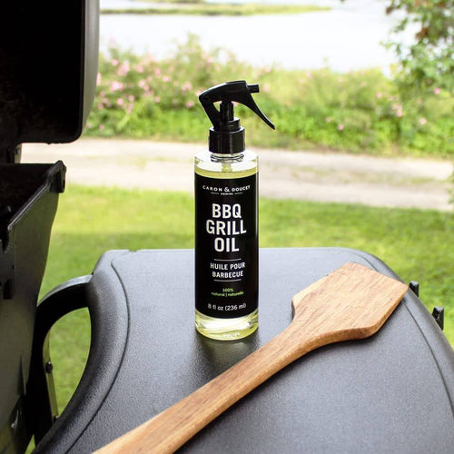 Clear spray bottle of natural BBQ grill oil cleaner with black label and wooden spatula resting on a black grill surface outdoors, showcased for home and grilling use.