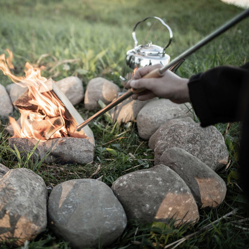 Person holding a long metal fire blow pipe with a wooden handle, directing air into a campfire surrounded by stones, set outdoors with a kettle in the background, ideal for Farm, Garden & Tool and Camping collections.