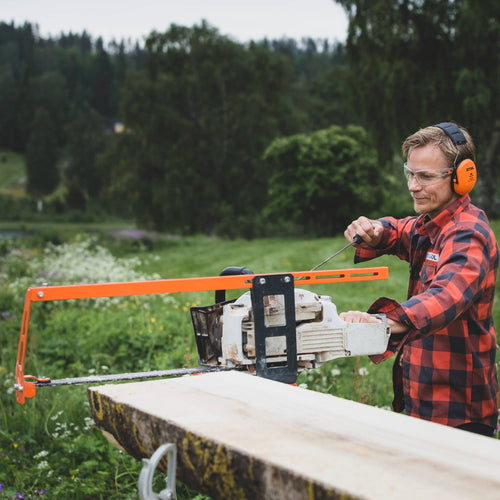 Person wearing protective earmuffs and goggles operating an orange bar nose steering accessory on a chainsawmill to align the guide bar for precise wood cutting in a green outdoor setting, featured in Farm, Garden & Tool and Wood Cutting and Hauling collections.