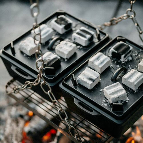Handcrafted cast iron loaf pans with fitted lids, heated over glowing coals on a suspended grill, ideal for outdoor cooking and baking in camping, kitchen, farm, and cast iron collections.