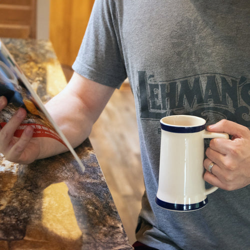 Person wearing a casual gray Lehman’s t-shirt holding a handcrafted stoneware stein mug with navy blue bands while reading a magazine at a kitchen counter, featured for Kitchen & Food, Dinnerware, USA Made, Best Sellers, and Promo Eligible collections.
