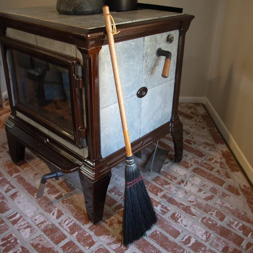 Handcrafted Amish-made broom with black corn straw bristles and natural wooden handle, leaning against a traditional wood stove, part of Hearth Accessories and USA Made collections.