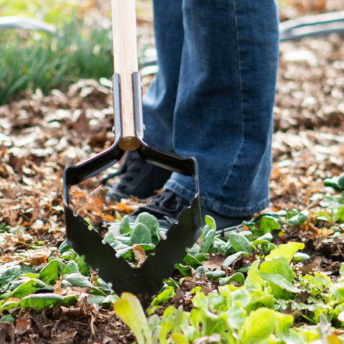 Handcrafted push-pull garden hoe with a long ash handle and serrated hardened steel blade being used to clear weeds among leafy green plants, part of USA Made farm and garden tools.