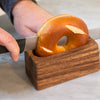 Handcrafted acacia hardwood bagel miter with wide base holding a bagel securely while a serrated knife slices through it, perfect for Kitchen & Food and Bread Making Supplies collections.