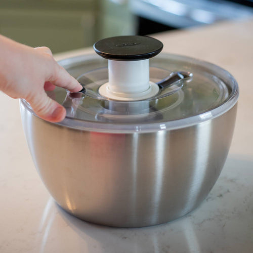Hand pressing the push-button handle to spin a brushed stainless steel salad spinner with locking clear lid and rubber bottom, featured in Kitchen & Food and Best Sellers collections.