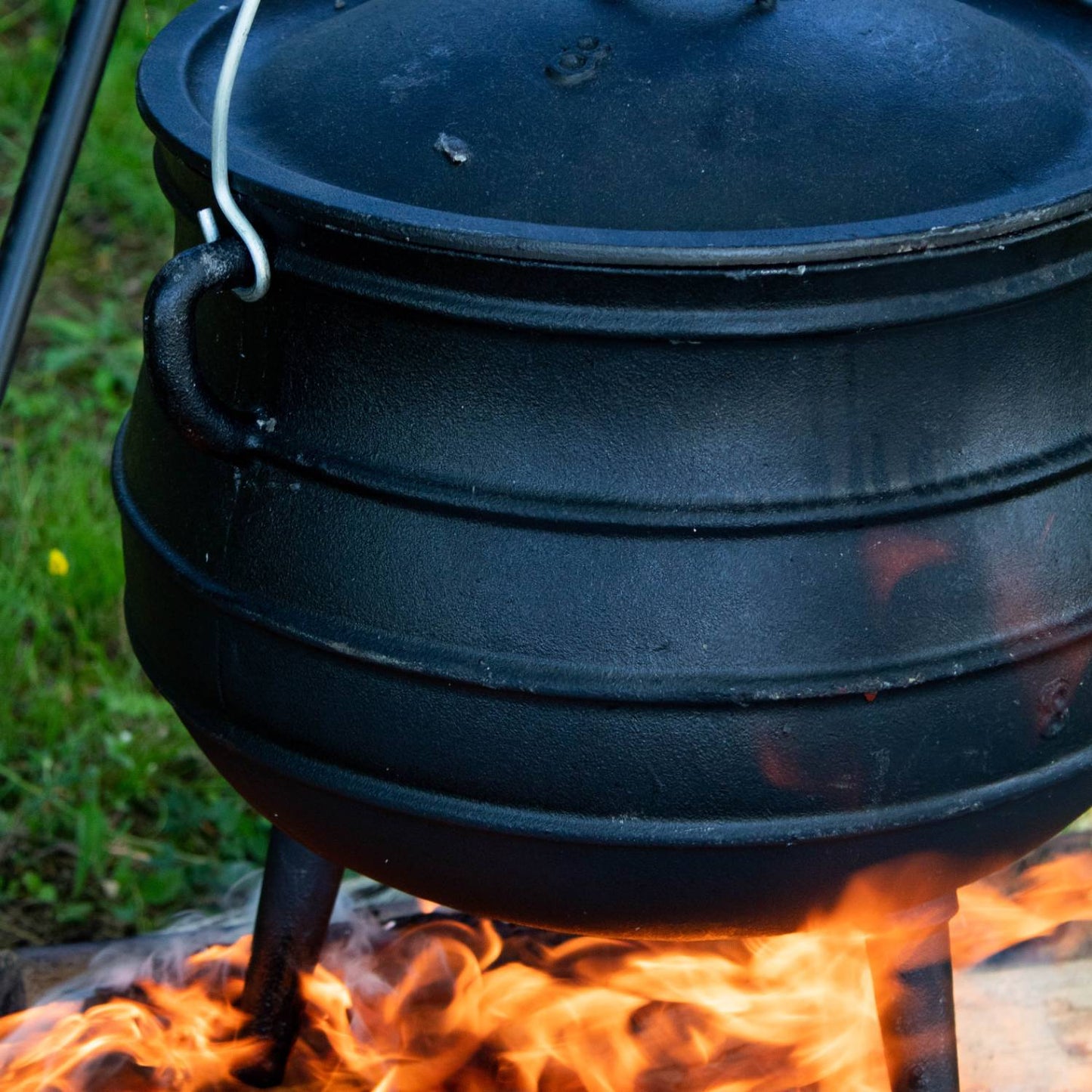 Close-up of a unique handcrafted cast iron kettle with a ribbed round body and three sturdy legs, sitting over an open flame, perfect for Farm, Garden & Tool, Camping, and Best Sellers collections.