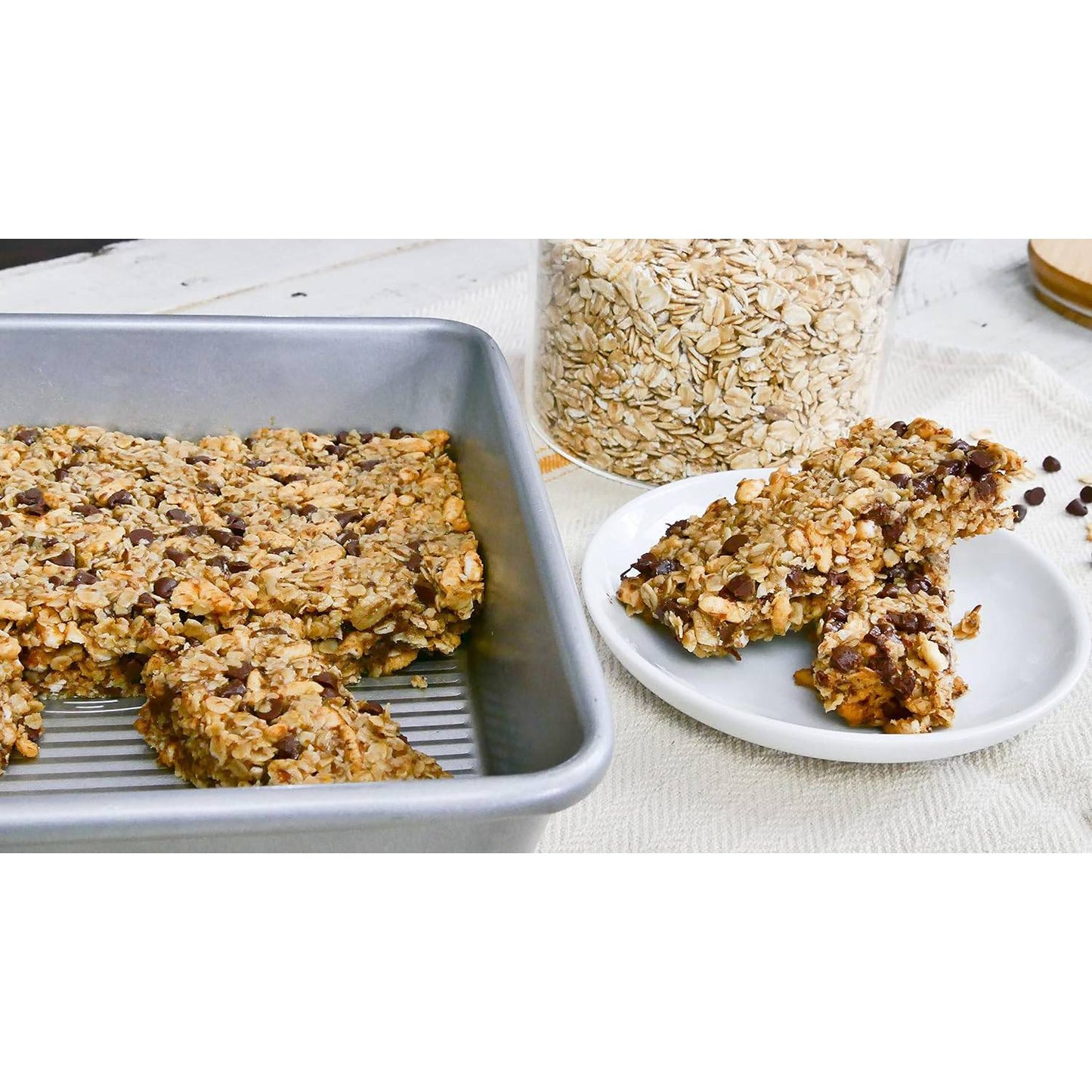 Aluminized steel baking pan holding freshly baked oat and chocolate chip bars with a few bars served on a white plate, styled in a kitchen setting with a jar of oats nearby, fitting Kitchen & Food and Baking Supplies collections.