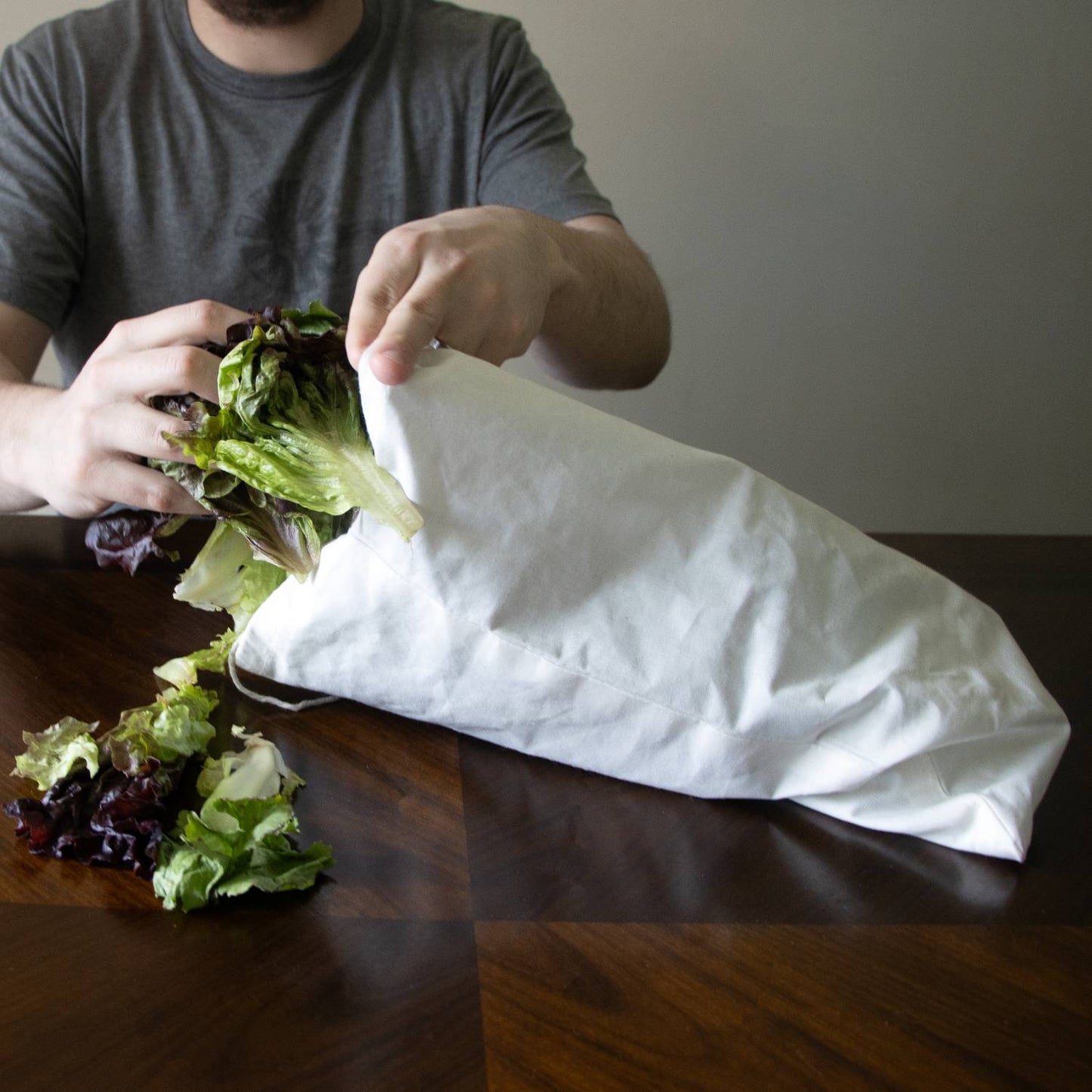 Person placing fresh lettuce into a white reusable drawstring bag made of breathable material, designed to keep greens crisp longer, featured in Kitchen & Food and Storage and Serving collections.