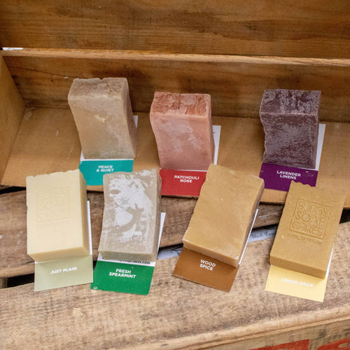 Seven handcrafted natural soap bars in assorted earthy tones with individual scent labels, displayed on rustic wooden shelves, highlighting Health & Wellness, Soaps, USA Made, and Best Sellers collections.