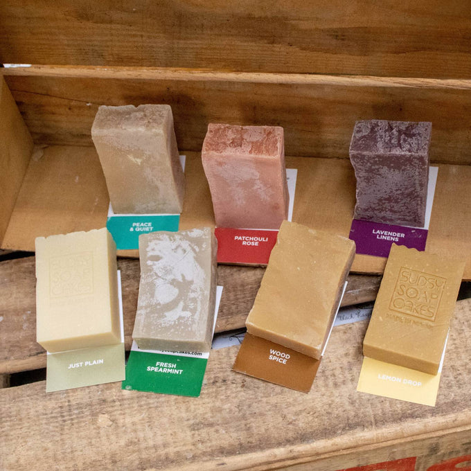 Seven handcrafted natural soap bars in assorted earthy tones with individual scent labels, displayed on rustic wooden shelves, highlighting Health & Wellness, Soaps, USA Made, and Best Sellers collections.