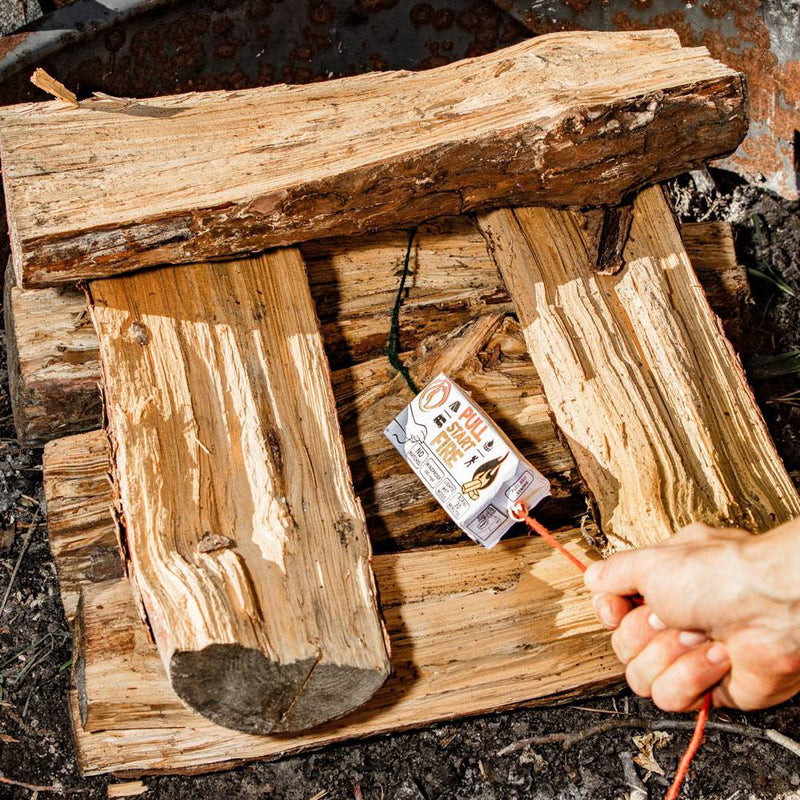 Hand pulling string fire starter ignited among split wood logs on ground, designed for reliable camping, farm, and garden use in any weather conditions.